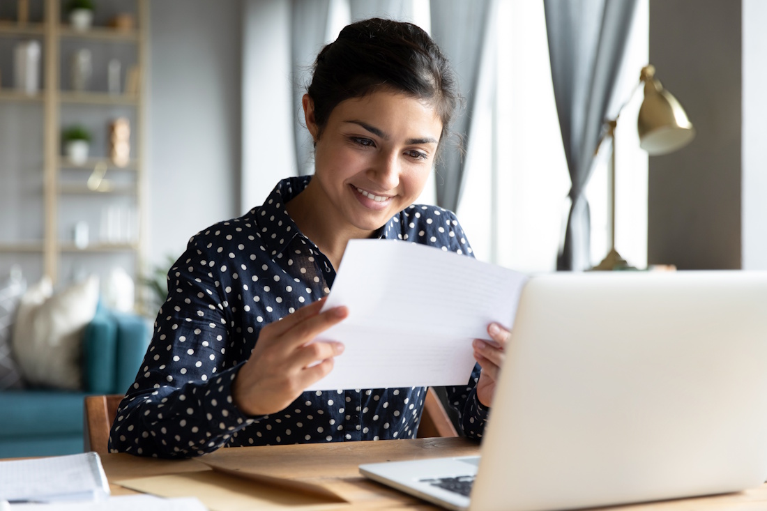A dark-haired woman in a blue long-sleeved polka-dot blouse sits at a table in a chic living space and smiles while reviewing a paper document, her laptop open on the table in front of her.