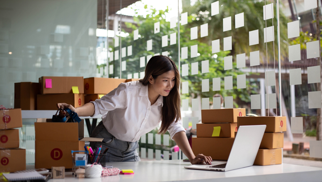 Image Alt Text	A young business owner manages online orders, checking her laptop while handling products and shipping boxes in a bright workspace.