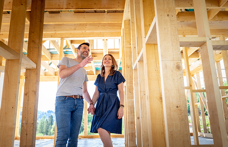 Couple standing inside unfinished home that has been framed. Couple is holding hands and smiling.