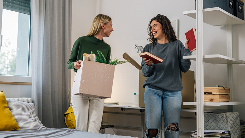 Two college aged women smiling and hanging out in dorm room.