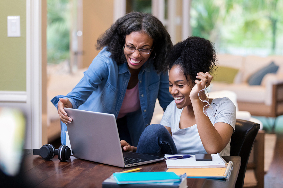 At a dark wooden table in an airy green and tan living space, a young woman in a pale blue t-shirt and dark jeans excitedly uses a silver laptop while a beaming older woman in a pink blouse and denim button-up looks on.