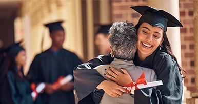 A smiling graduate in a cap and gown hugs an older woman, holding a diploma tied with a red ribbon.