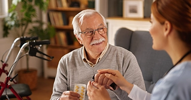 A smiling elderly man with glasses and a walker receives medication from a caregiver in a home setting.