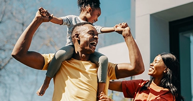 A Black father carries his daughter on his shoulders while his wife smiles beside them, all outdoors.